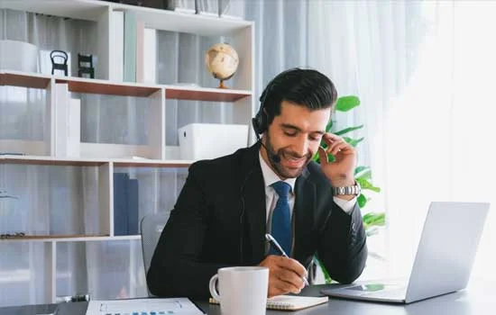 A businessman in a suit, wearing a headset, smiles while writing notes at a desk with a laptop and coffee mug. The office background is bright and organized while symbolizing a supporting