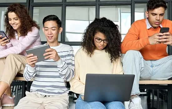A group of four young adults seated, engaged with devices phones, a tablet, and a laptop, in a bright room. The mood is casual and focused.