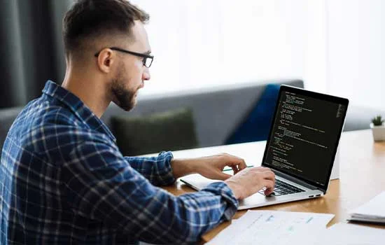 A man in a plaid shirt sits at a table, focused on writing code on a laptop. The setting is a modern living room, conveying a calm, productive mood.