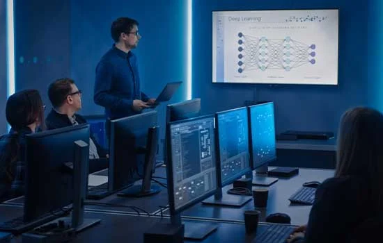 A man presents a deep learning diagram on a screen to colleagues seated at desks with computers. The room's blue lighting conveys a tech-focused atmosphere.