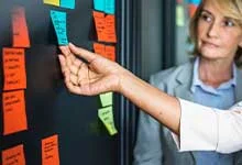 A person places a blue sticky note on a board filled with colorful sticky notes. Another person observes, suggesting teamwork and planning in a professional setting.