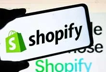 A phone displaying the Shopify logo, featuring a green shopping bag icon and black text, is held up against a blurred background with similar text.