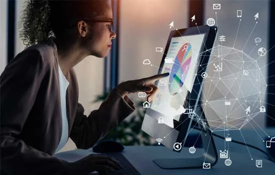 A woman in a dimly lit office interacts with a glowing computer screen displaying graphs. Transparent digital icons and a network sphere overlay the scene, suggesting data analysis and technology.