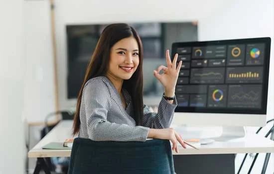 An asian woman in an office setting gives an OK hand gesture while smiling at the camera. She sits at a desk with a computer displaying graphs, conveying confidence.