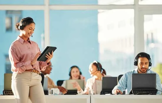 An office scene with a smiling woman teal and light brown pants using a tablet, while four colleagues with headsets work at laptops, creating a collaborative atmosphere.