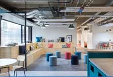Modern office space with open layout, featuring gray and blue cushioned stools, wooden benches with colorful pillows, and industrial-style ceilings in a workspace.