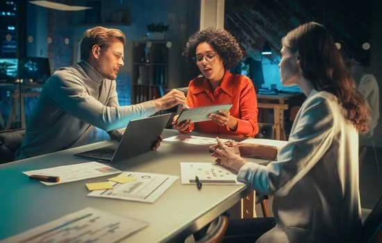 Three colleagues collaborate in a modern office, engaged in discussion around a tablet and laptop, with documents on the table, creating a focused atmosphere.