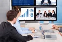 Three people in an office have a video conference. Two screens show business charts and a meeting with colleagues, conveying professionalism and focus.