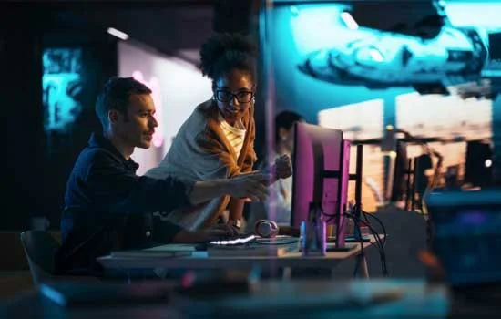 Two people are collaborating at a computer in a dimly lit office. They look focused and engaged, with a large glowing screen and modern workspace.
