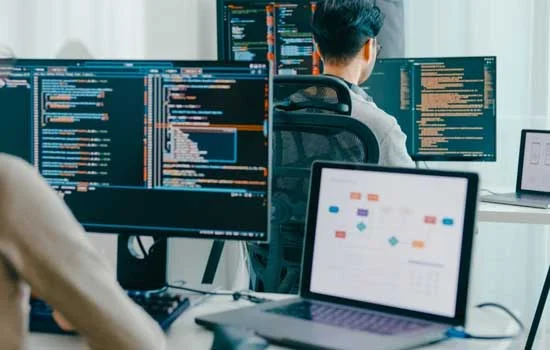 Two people are working at computers with multiple screens displaying code and flowcharts. The scene conveys focus and collaboration in a tech environment.