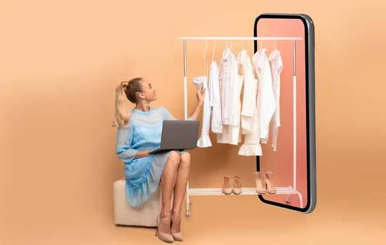 Woman in blue dress sits with a laptop, facing a large smartphone displaying a rack of white clothes and shoes, against a peach background. Embracing online shopping.