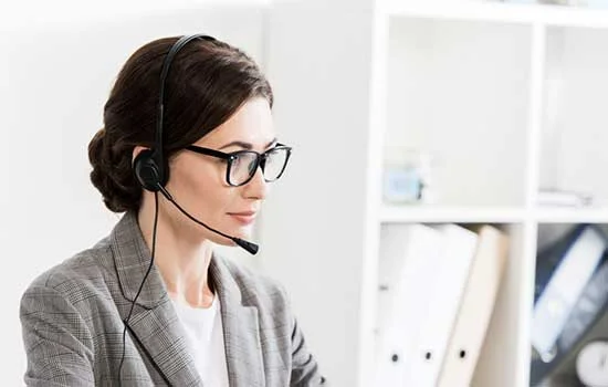 Woman in glasses with a headset, wearing a gray blazer, working in an office. Focused expression, bookshelves in the background. Professional ambiance.